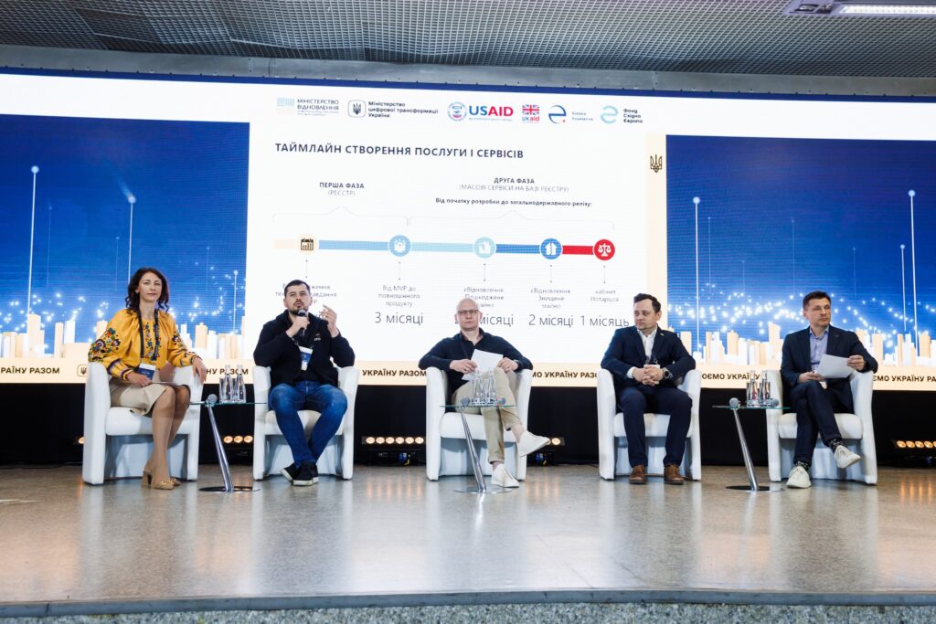 Five people sit on stage in front of a large screen wall. The screen displays information about eRecovery and the logos of USAID, UK Aid, Ukraine's MCTID, MDT, Eurasia Foundation, and the East Europe Foundation.