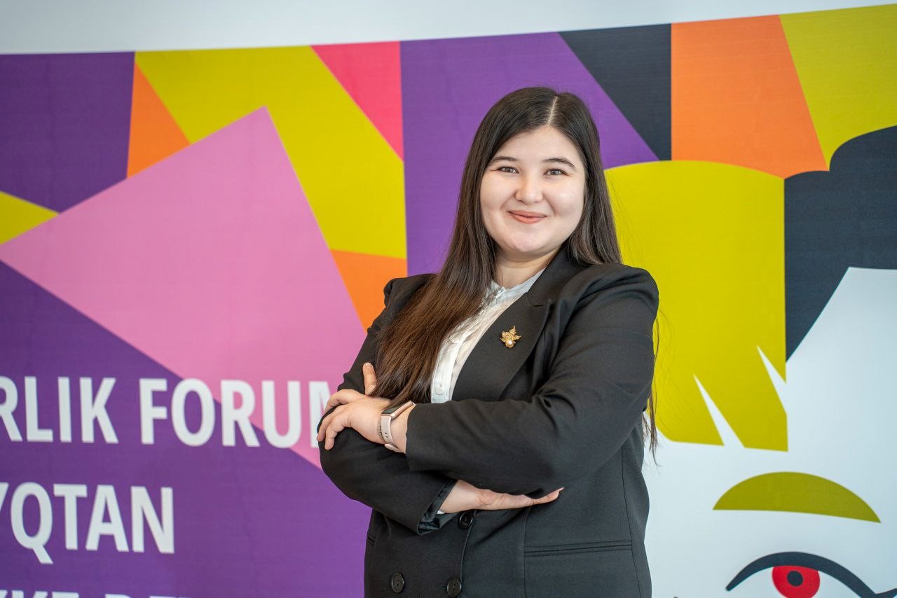 Nasiba, a young woman from Kazakhstan, crosses her arms and smiles confidently into the camera. She posed in front of a brightly colored banner for a forum.