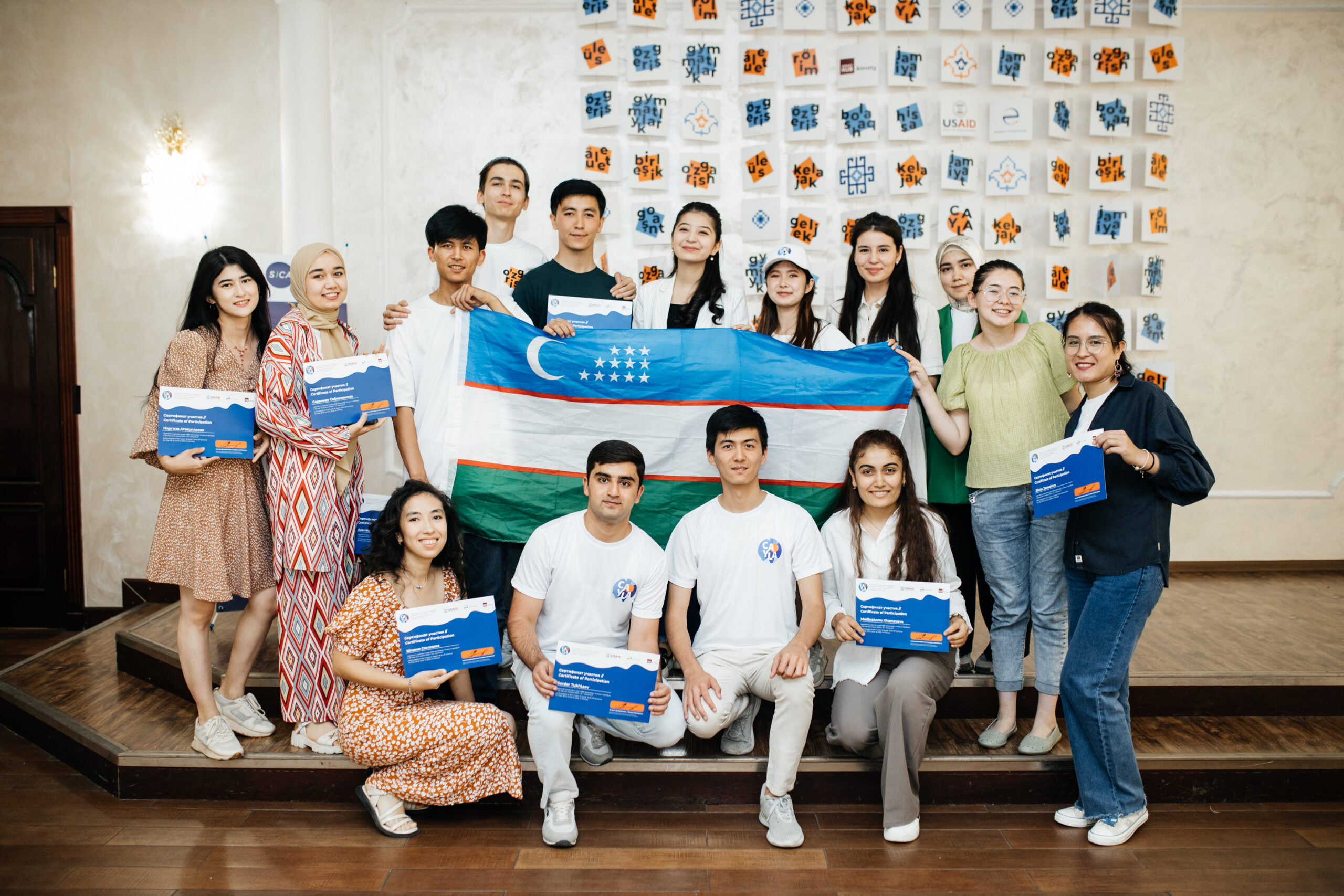 Fifteen young leaders from Uzbekistan pose with their CAYLA certificates of completion and a large flag of Uzbekistan.