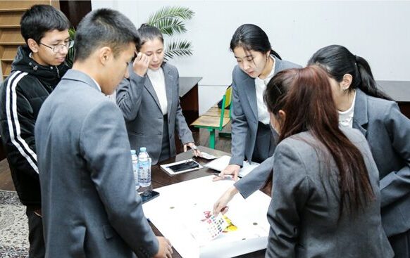 A group of 6 teens in school uniforms gathers around a table to complete a learning activity together.