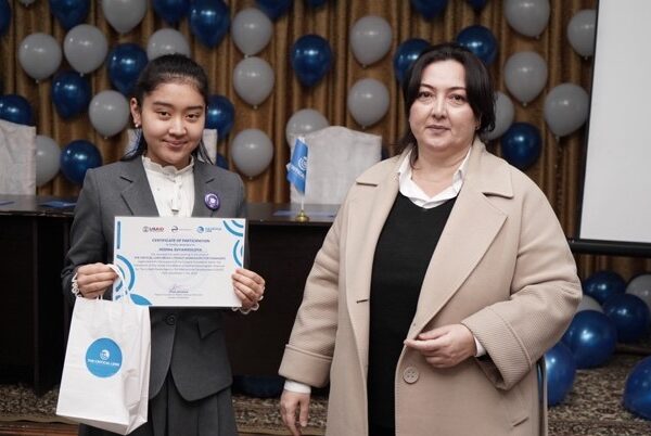 A teenage girl and a teacher stand in a room with blue and white balloons on the wall. The girl smiles and holds up a certificate of completion.