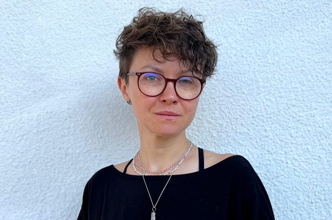 A portrait photograph of Alesya Nugayeva. She stands facing the camera, angled slightly to the left. She wears glasses and a black shirt.