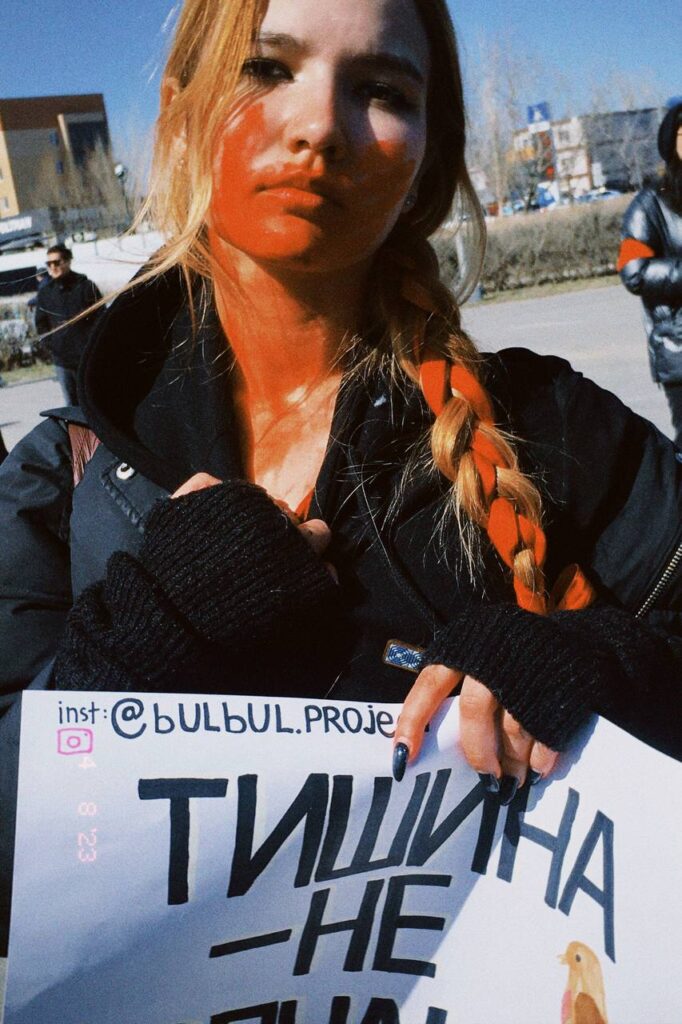 Alina Ilnitskaya stands close to the camera holding a poster. Most of the text is obscured except one word--"Silence." The poster includes the Bulbul Instagram handle and an illustration of a bird. Alina has red paint smeared on her face and in her hair, presumably to resemble blood.