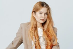 Alina Ilnitskaya sits on the arm of a white recliner against a white background. Her body is angled to the right. She is wearing a tan blazer, white tee, and jeans, and her hair hangs in loose curls. She is looking directly at the camera. Her expression is determined.