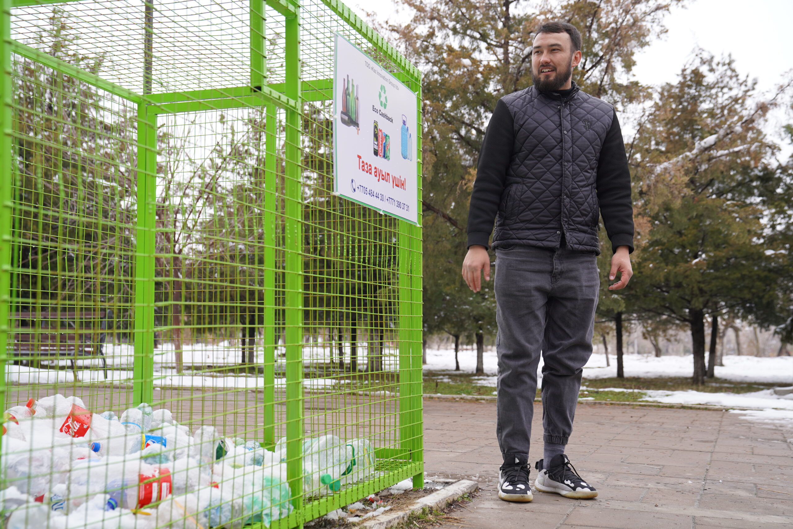 Vladislav stands to the right of a green recycling collection container. He is looking at the container sign and smiling. He is wearing a black shirt, black quilted vest, black jeans, and black shoes. There is snow on the ground behind him.