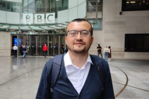 Mirzo is photographed standing outside the entrance of the BBC Broadcasting House headquarters in London. He is wearing glasses, a white button-down shirt, dark blue cardigan sweater, and a backpack.