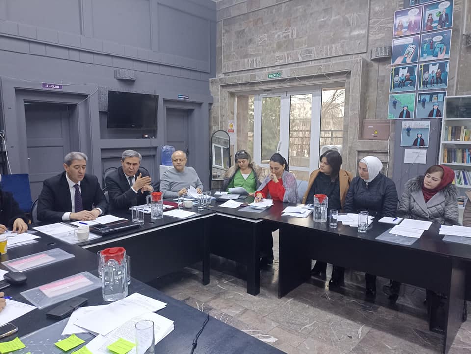 A roundtable discussion is taking place. Only eight of the people seated at the table are photographed. In the back section of the table are three men, and seated perpendicular to them are five women. The women occupy the right side of the photograph. The man photographed in the center of the three is talking to someone to his side not in the photograph. His hands are open-palmed. One crossed atop the other. Everyone around him is listening closely.