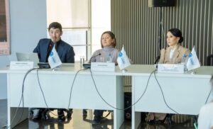 Three people are seated at a table. They are speaking at an event. A man, Mirkhat, is seated to the left in the image. Two colleagues, both women, are seated to the right of him.