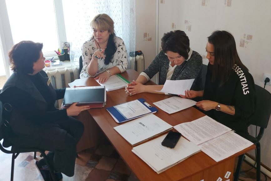 Four women from the Zhenskii Luch team are seated at a table working in two pairs. One pair is talking, while the other pair is looking over documents that are on the table.