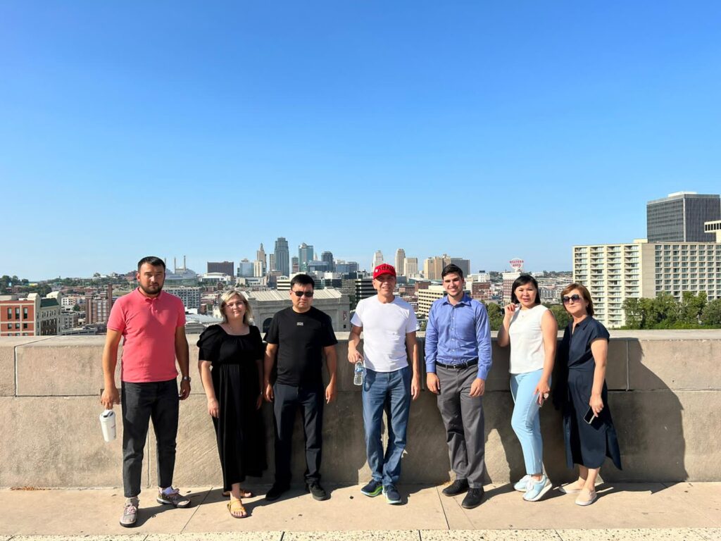 A group of seven individuals stands in front of a concrete barrier, with a cityscape behind them.