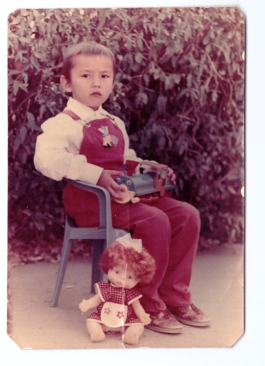 A childhood photo of Vladislav Golyarko. A little boy in red overalls sits in a chair holding a toy train, with a doll at his feet.